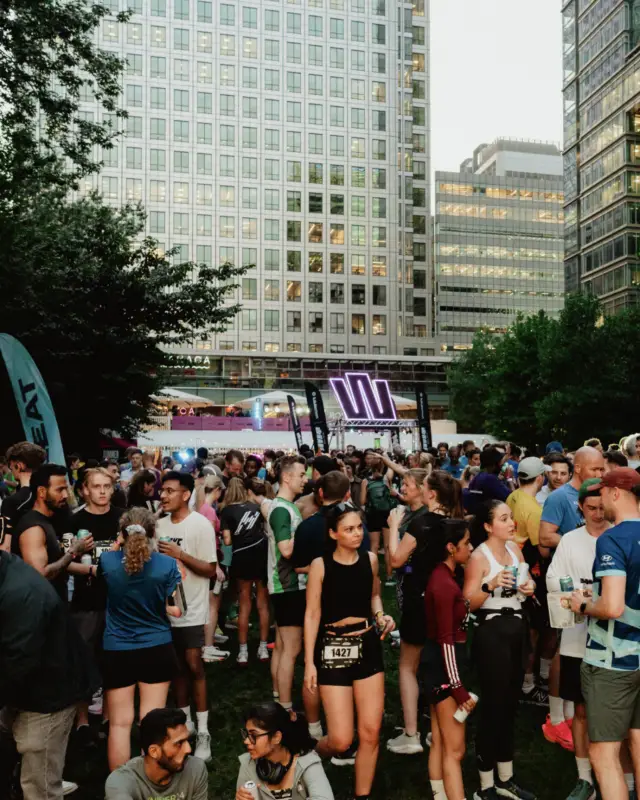 The Event Village crowd at dusk with the neon W stage glowing under One Canada Square